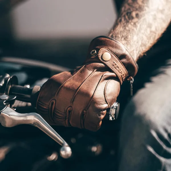 Rider wearing brown leather motorcycle gloves with reinforced knuckles and secure snap button closure, gripping a motorcycle handlebar for a rugged and stylish look