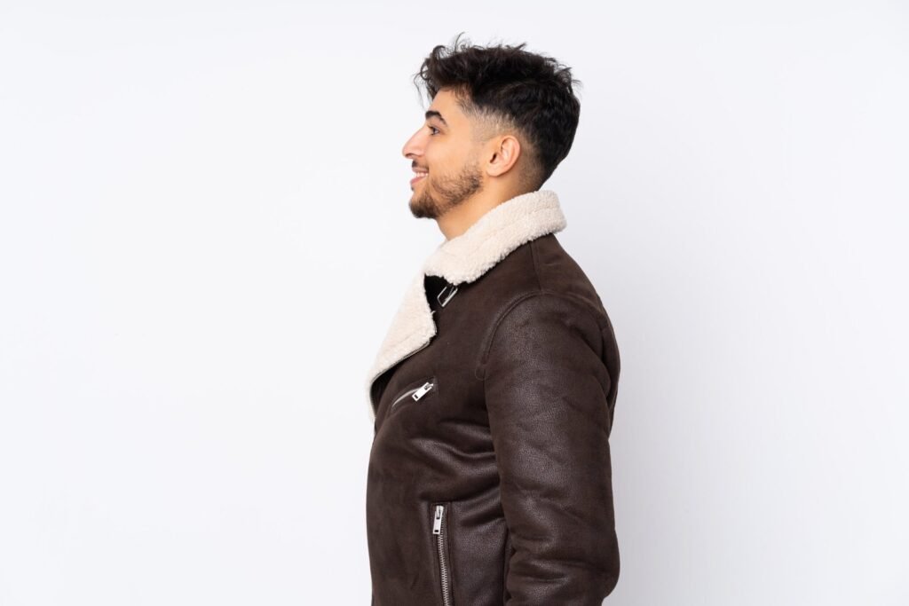 Side profile of a smiling young man wearing a brown shearling leather jacket, set against a clean white background