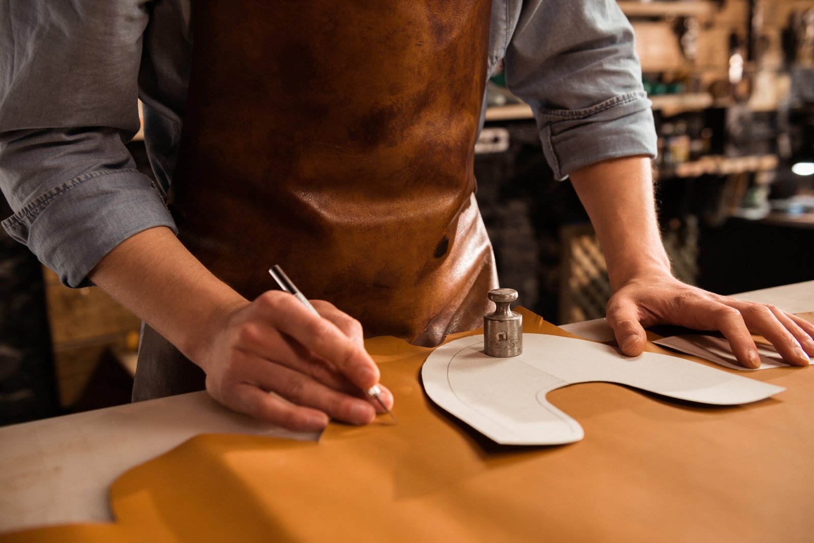 Craftsman wearing a leather apron, carefully tracing a pattern onto a piece of leather with a precision knife in a workshop setting